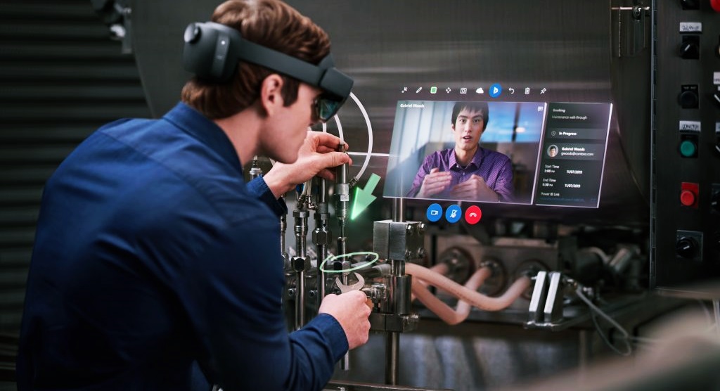 Un hombre trabaja a distancia viendo a otra persona en un monitor y con lentes para realidad aumentada 